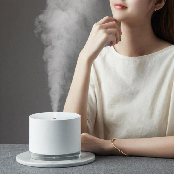 Person using a white Beau Glow  Humidifier & Ambient Lamp with steam rising, sitting on a gray surface.