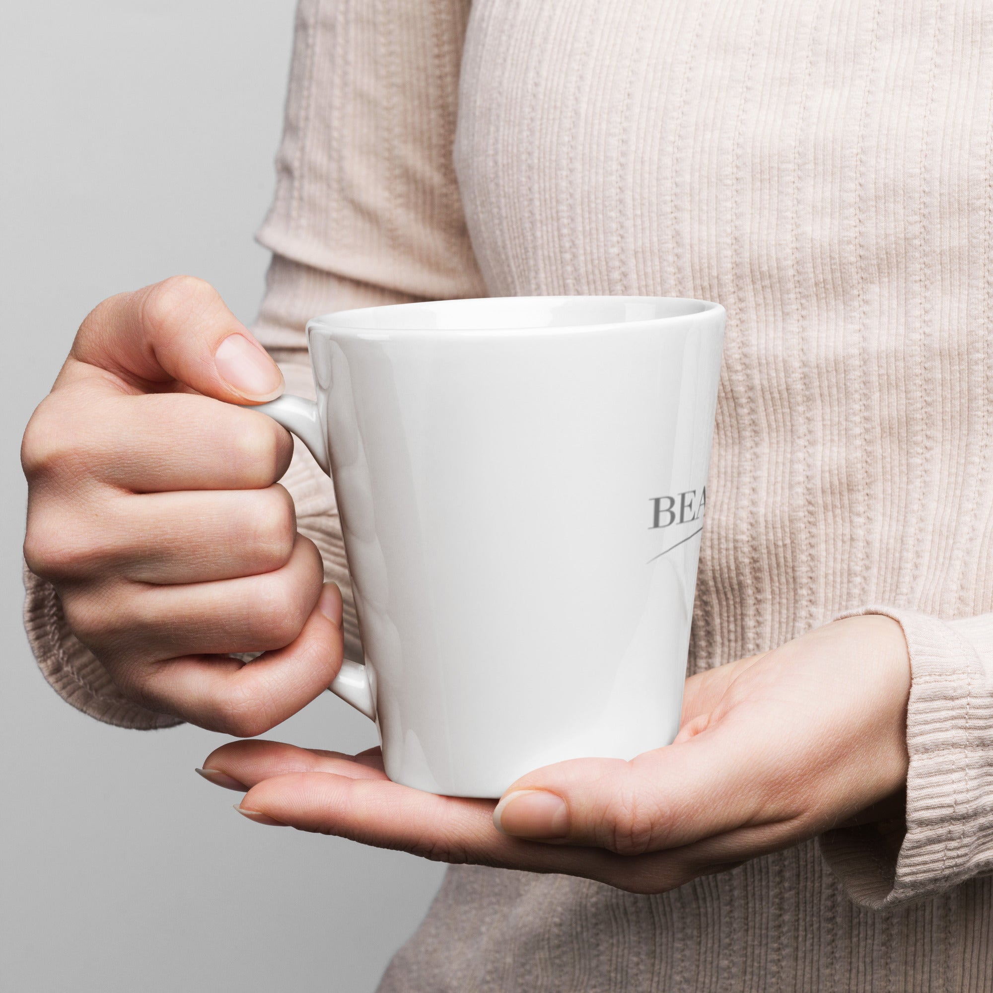 A close-up view of a person wearing a soft knit sweater holding the white ceramic Beau House latte mug with both hands.⁠