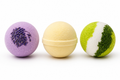 
Three handcrafted round bath bombs lined up on a white background: a purple lavender bomb with dried flower buds, a smooth pastel yellow lemongrass bomb, and a vibrant green and white eucalyptus bomb.
