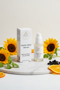 BEAU  Wellness & Beauty Hydra-Glow Serum shown with its white product box and clear pump bottle on a clean white background with sunflower., elderberries and watercress and orange slices.