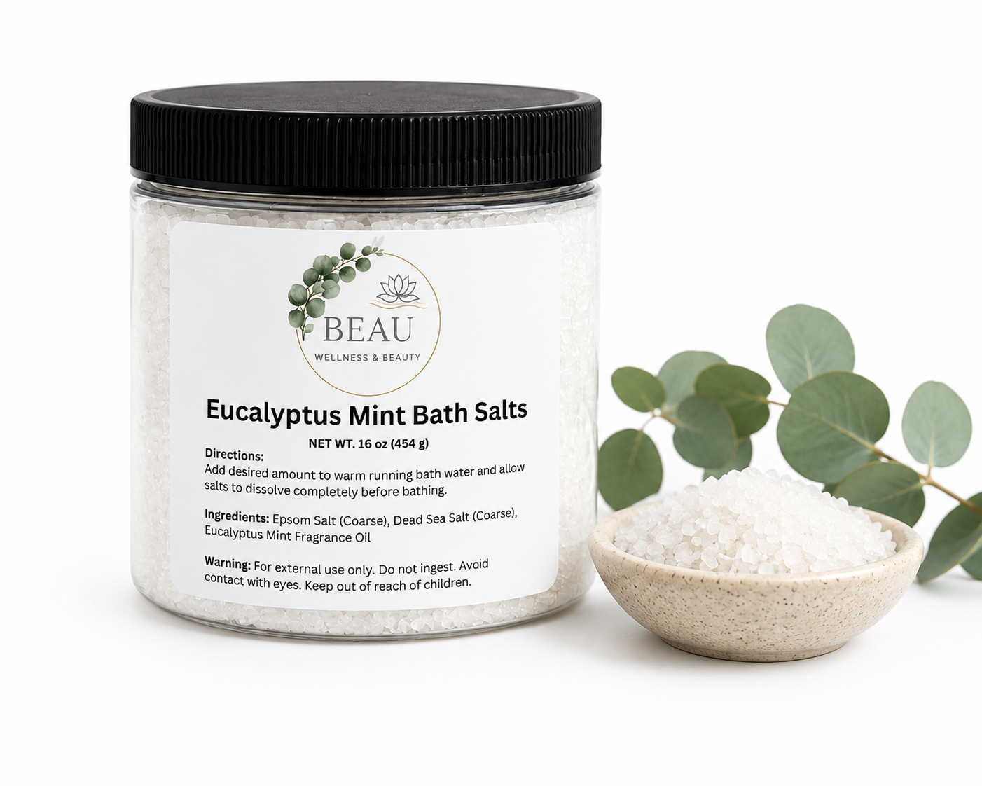 A clean, front-facing product shot of a clear jar of Eucalyptus Mint Bath Salts with a black lid and white label, sitting next to a small white bowl filled with bath salts and fresh green eucalyptus leaves on a white surface.